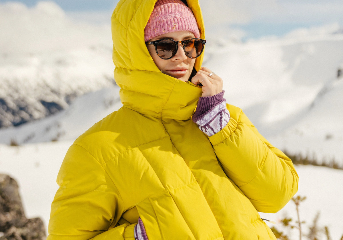 Femme dans un paysage hivernal montagneux portant une doudoune courte jaune FOSFO et une tuque rose. 