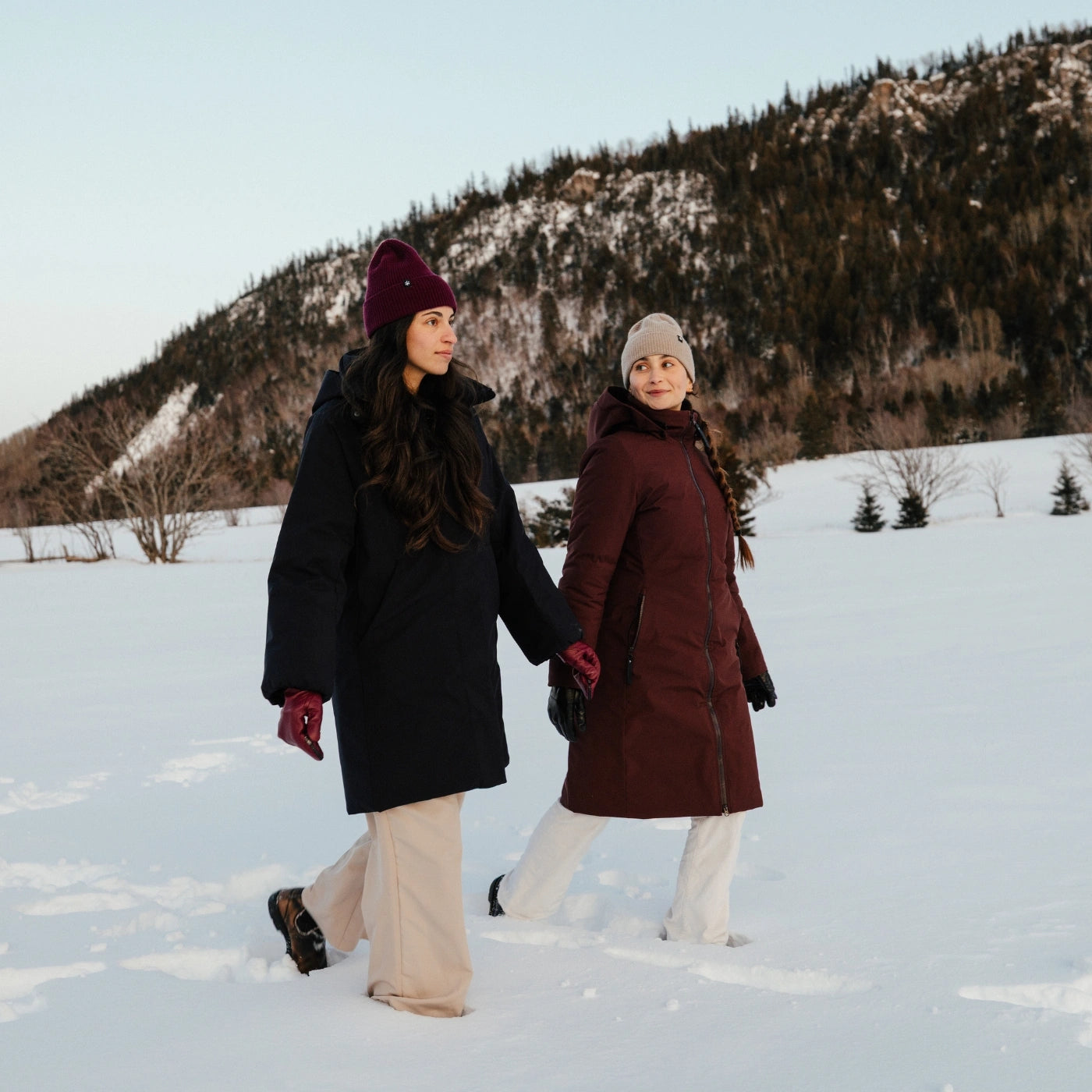 Deux femmes marchant dans la neige portant des manteaux longs d'hiver noir et bordeaux de la marque Audvik.