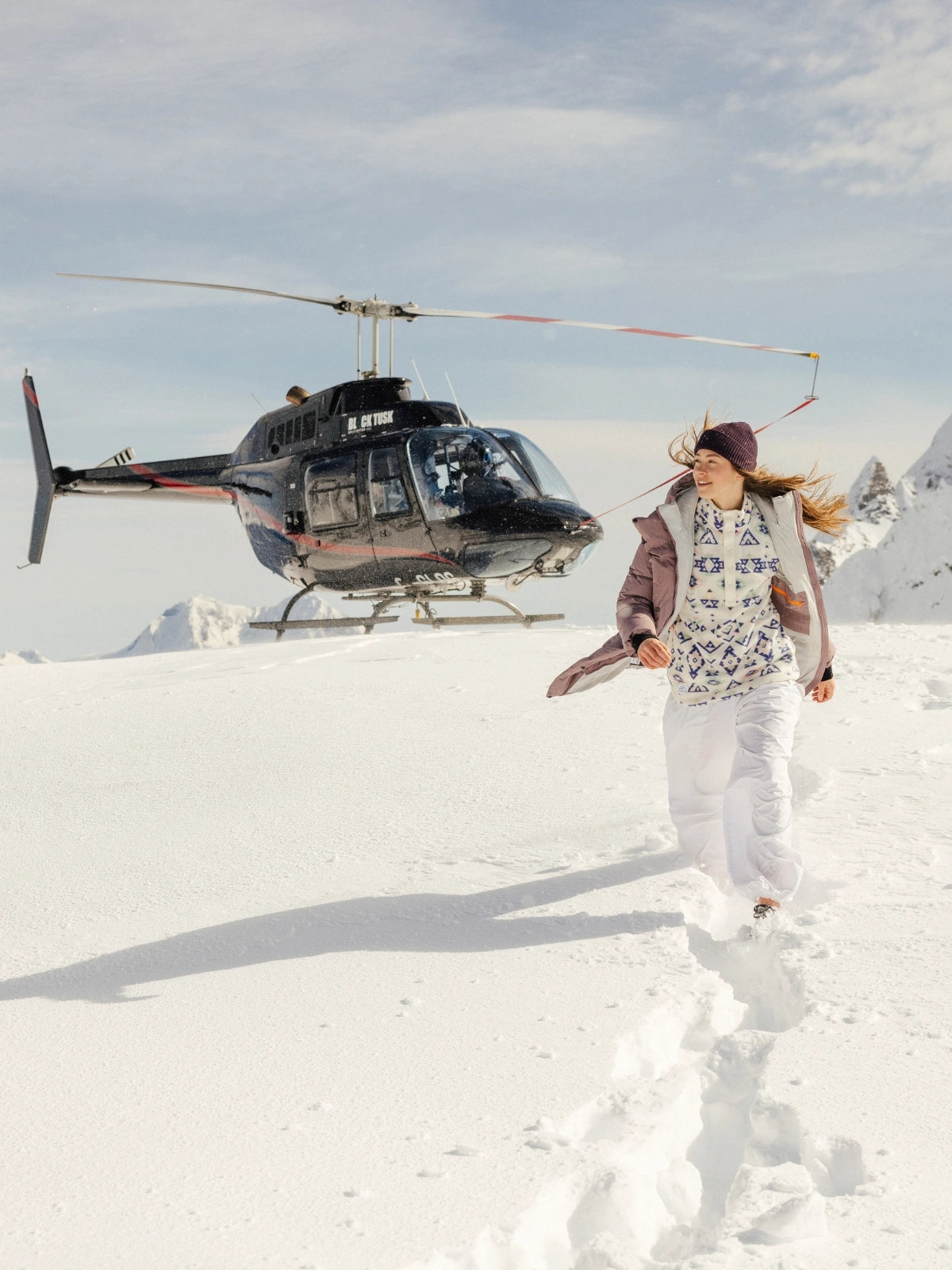 Femme dans la montagne enneigée marchant avec une doudoune FOSFO devant un hélicoptère. 