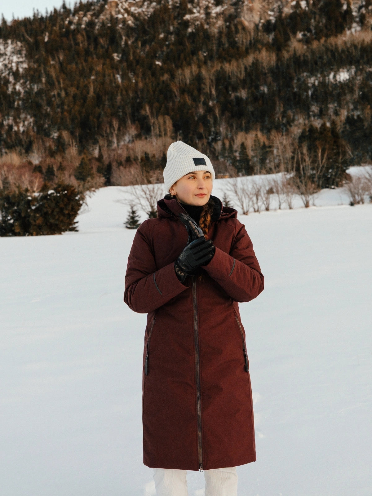 Femme dans un paysage hivernal portant un manteau d'hiver long de couleur bordeaux et une tuque beige Audvik.  