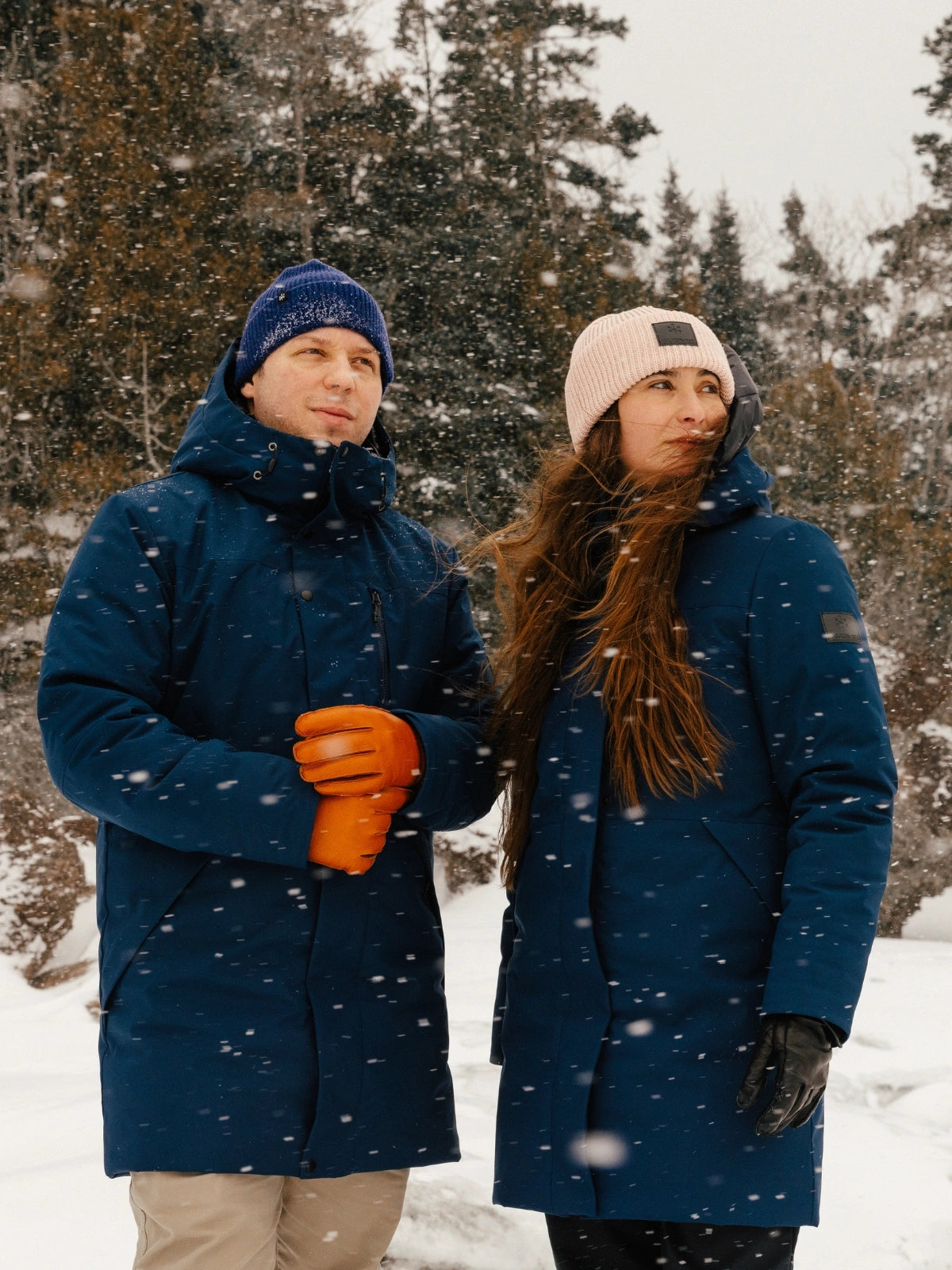 Un homme et une femme dans un paysage hivernal portant chacun un manteau d'hiver Audvik bleu marine et des tuques Audvik.