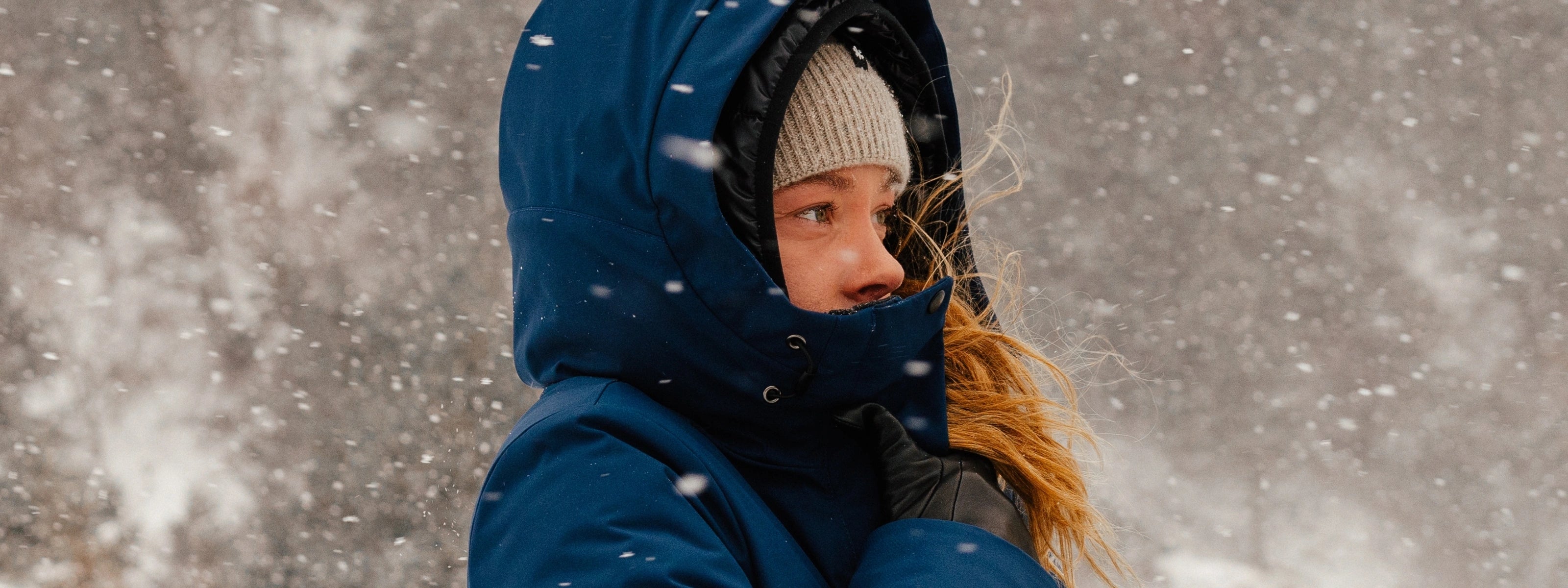 Femme dans une tempête de neige portant un manteau d'hiver marine Audvik. Elle a la capuche sur la tête ainsi qu'une tuque en cachemire Audvik beige. 