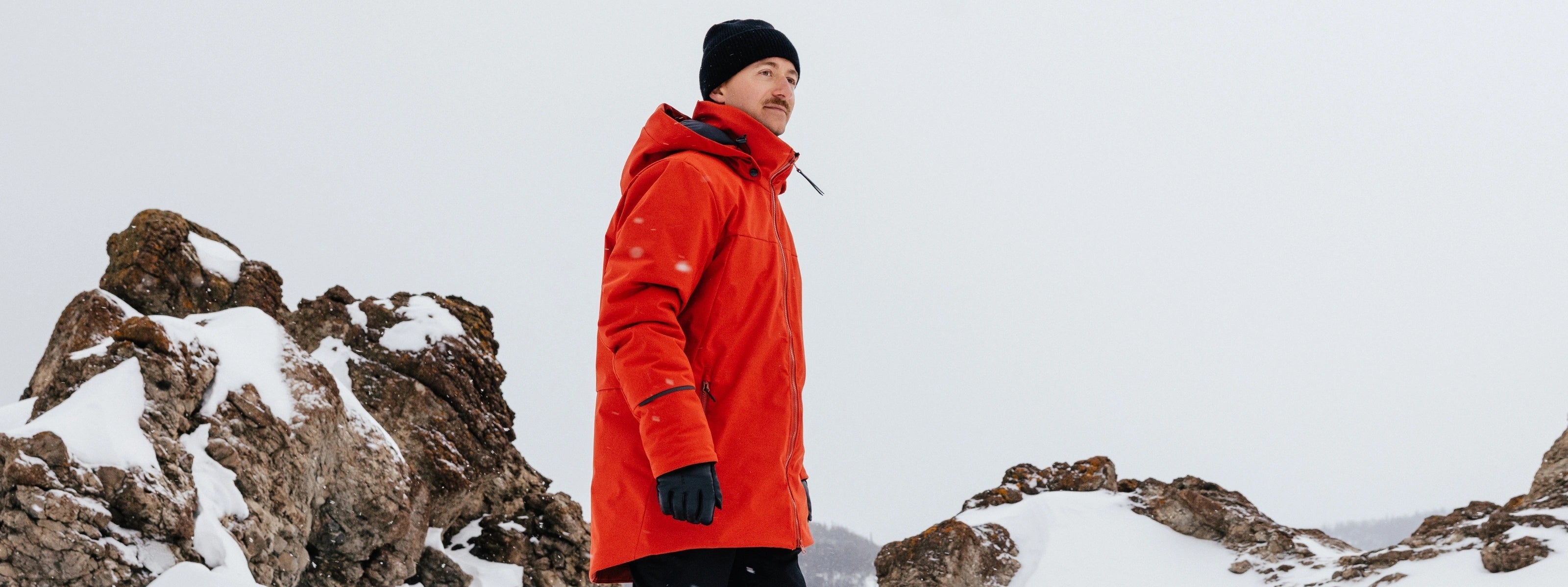 Homme dans un paysage rocheux enneigé portant un manteau d'hiver Audvik rouge et un bonne en cachemire noir